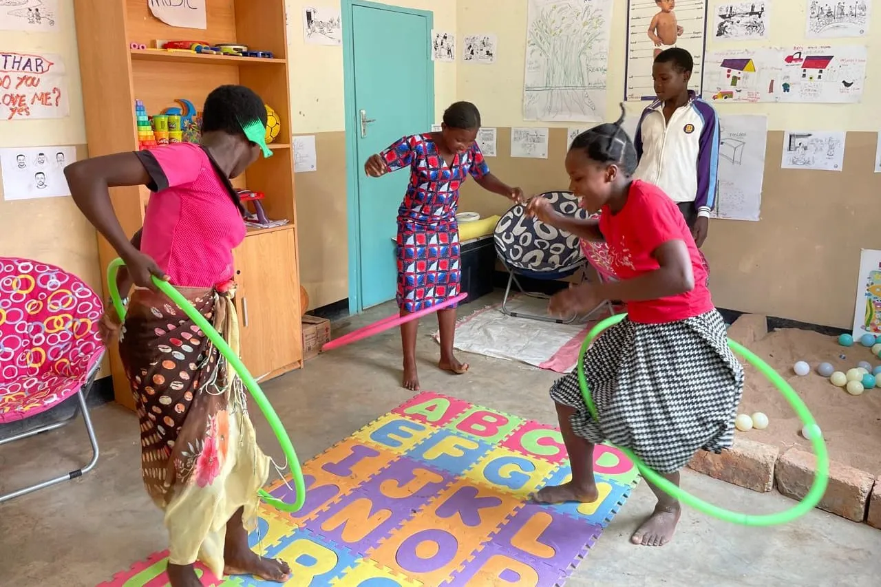 Young people swing hula hoops on an alphabet play mat in a room hung with children's drawings, at a group therapy and expressive arts session in Zambia