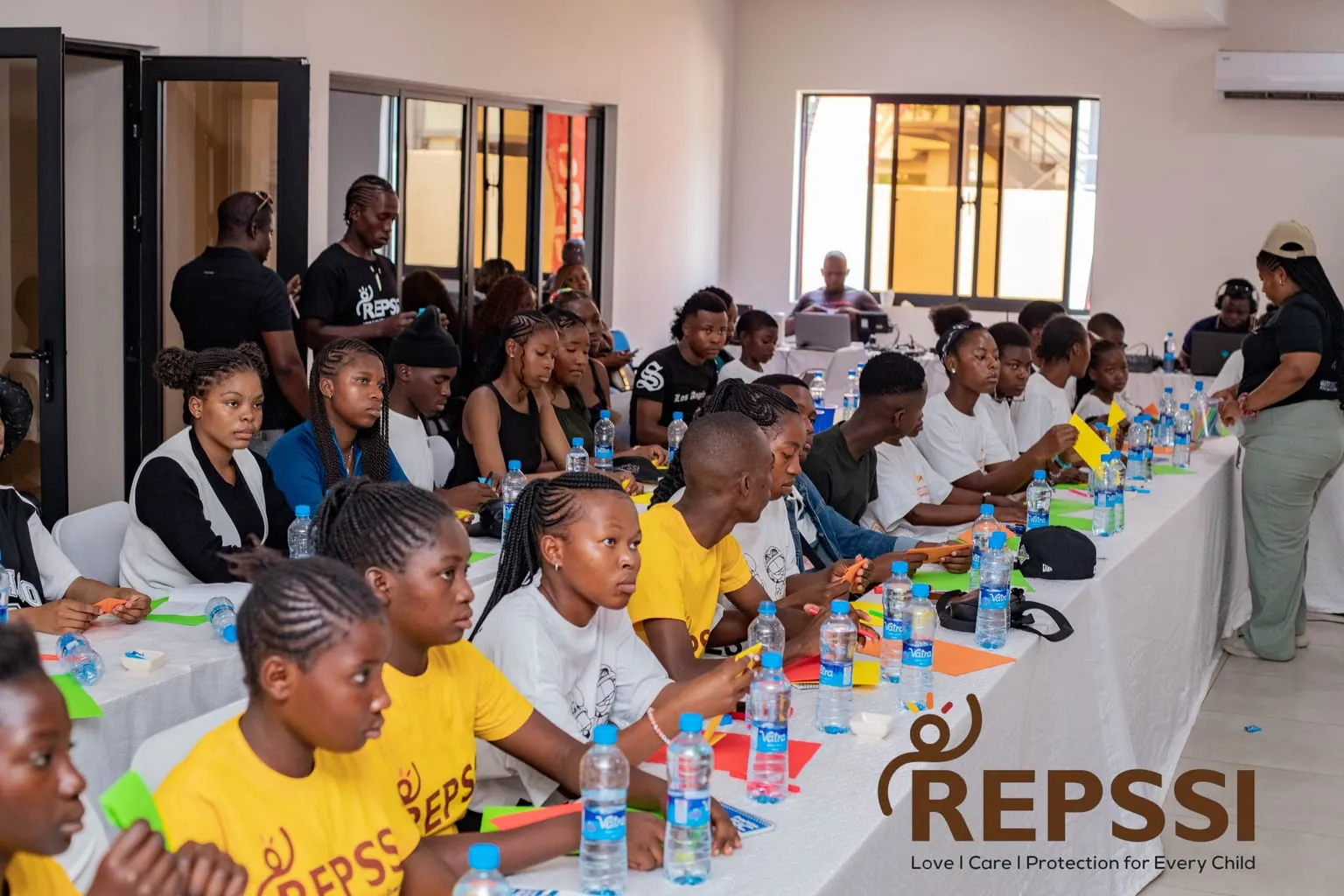 Young people, many in REPSSI T-shirts, sit along a long table with coloured paper and water bottles, listening during a workshop